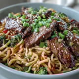 Steaming skillet of Korean Beef Noodles with bell peppers and carrots, garnished with green onions and sesame seeds.